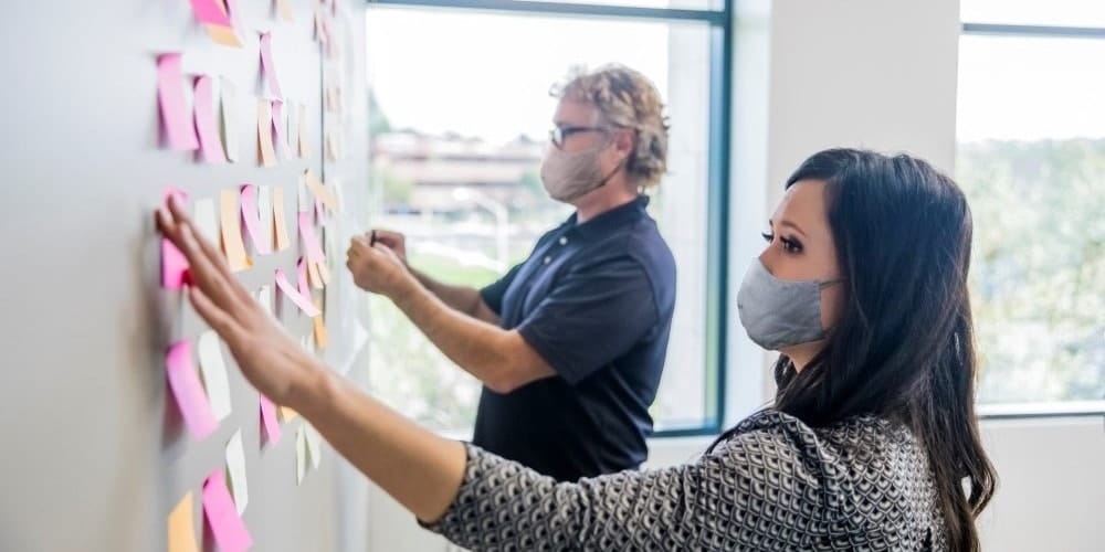 Two business people in masks pinning post-its to a wall